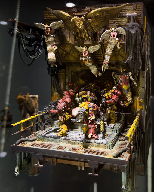 Photo of a miniature diorama, showing Howling Griffons marines in their red-and-yellow armour pushing out onto a walkway from a dark corridor.