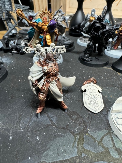 Photo of a Valius miniature on a painting table. The model has been undercoated an off-white colour and the armour painted a bronze-like colour. The model is off its base for painting, while the arm with shield is also separate, lying on the table.