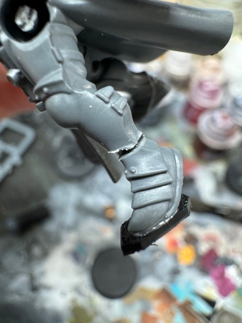 A close-up photograph of the lower leg of a plastic miniature. The foot has been pinned to the leg, with a gap showing between the pieces