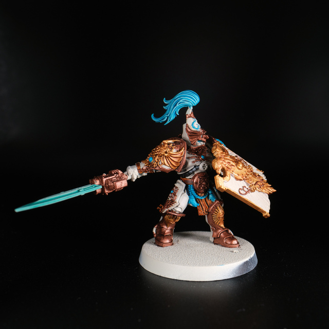 Photo of a lit miniature on a black background. An Adeptus Custodes figure with shield held up in front and sword arm trailing back behind it. The base armour colour is an off-white, with bronze and gold metallic paint to the detail and embellished areas.