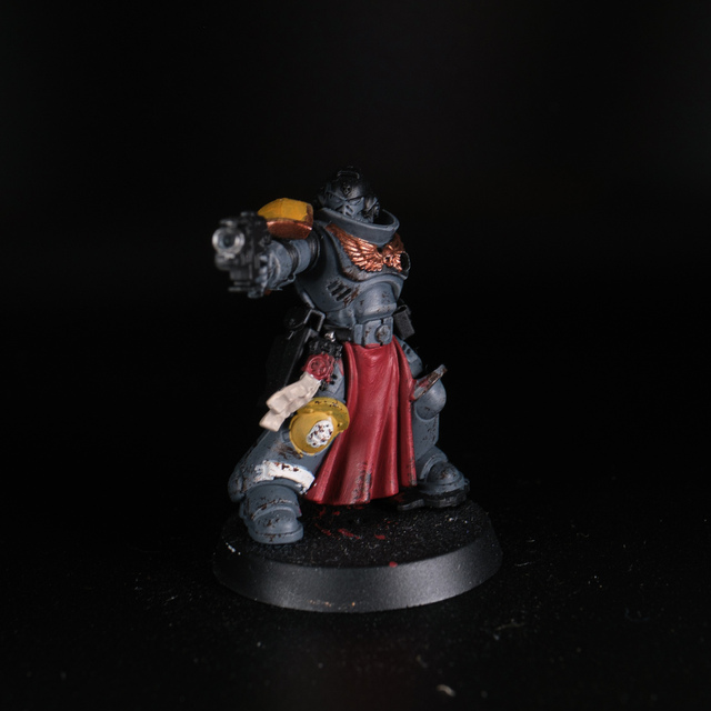 Photograph of a lit space marine miniature on a fully black background. It's unfinished, with an arm and the backpack missing and areas not painted yet, but has dark grey Space Wolves armour flecked with 'mud' and a long red loincloth hanging from its waist.