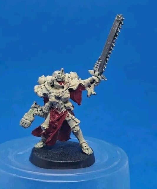 Low quality photo of a Sisters of Battle model on a blue background. The model is painted mostly in an off-white, with crimson robes and looks very blotchy