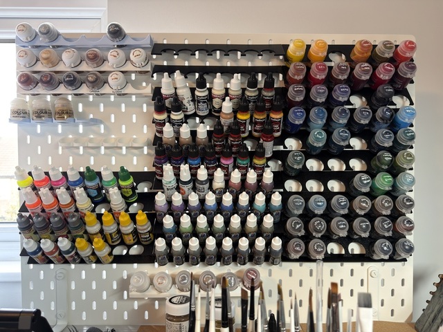 Photo of a white Ikea Skadis peg board with many black 3D-printed paint racks mostly filled with paints. Each 'shelf' angles steeply down away from the pegboard, with a series of circular holes cut out of the front edge to allow paint bottles to be held. Each paint bottle hangs in its shelf and the bottom rests on the back of the shelf below it.