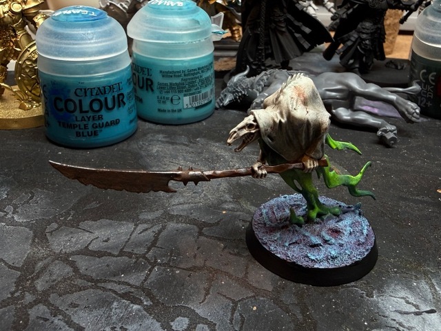 Photo of a painted Glaivewraith Stalker model on a painting table. It has dirty pale rags and bright green glowing trails, on a base that has magenta shadows and bright pale blue edge highlights