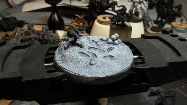 Photo of a large sculpted miniature base on the XL painting handle from Citadel, showing a snowy ground with footprints and some snowballs piled on one side. The snow has been painted, with a pale blue-grey undertone and white catching across all the highlights for a snow effect