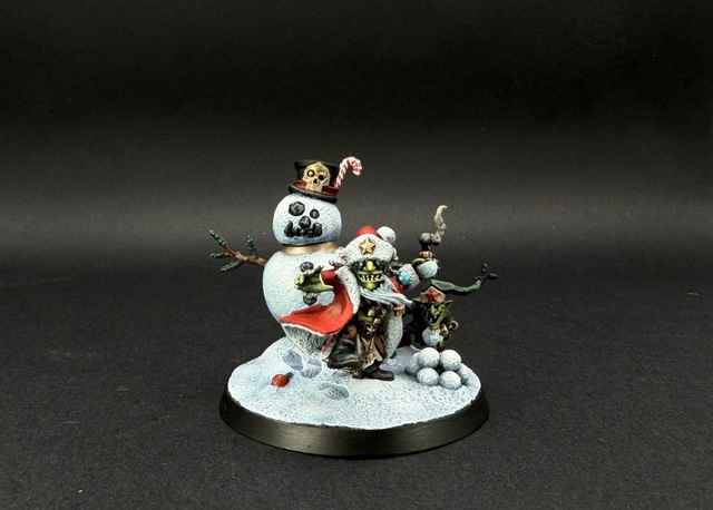 Photo of the fully painted miniature, on a black backdrop. Da Red Gobbo is in a red coat with white fleece lining, while the gretchin elf sneers out from behind him in a dark green outfit. The snowman’s head just shows a bit of a warm metallic ‘neck’ beneath.
