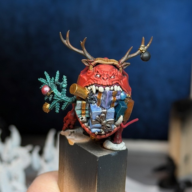 Photo of a squig model tacked to a wood block, with some painting started. The squig has red flesh, with highlights added to all the wrinkles. The presents in its mouth have pale coloured-metallic paints down that don’t quite work yet.