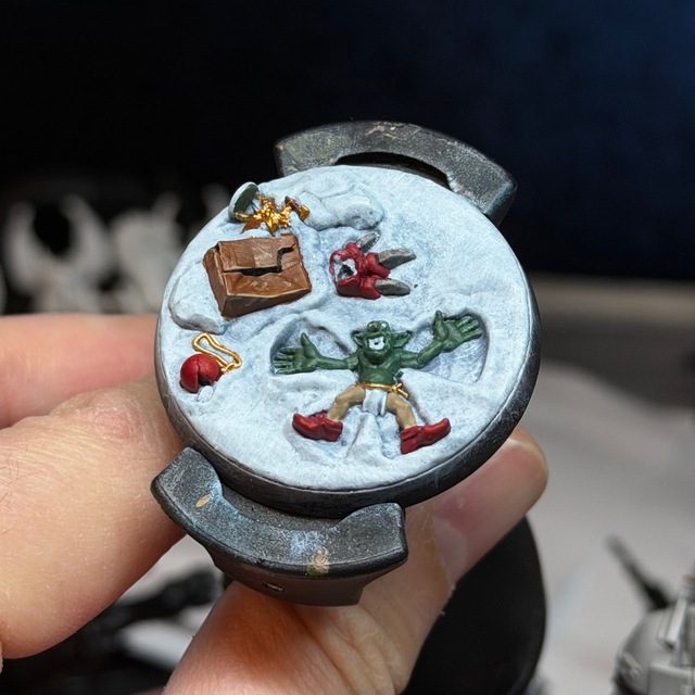 Photo of the base on a painting handle. There’s a grot making snow angels in the snow, a brown crumpled box, a bauble, a gold-painted Stormcast Eternal miniature on its base and the squig’s standing foot
