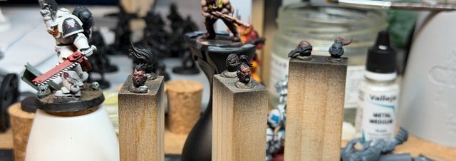 Wide ratio view close in on three wooden blocks with space marine heads stuck in white tack. Most are bare heads with one helmet and each of the bare heads has at least a base coat of paint to the skin, with simple highlights on a couple.
