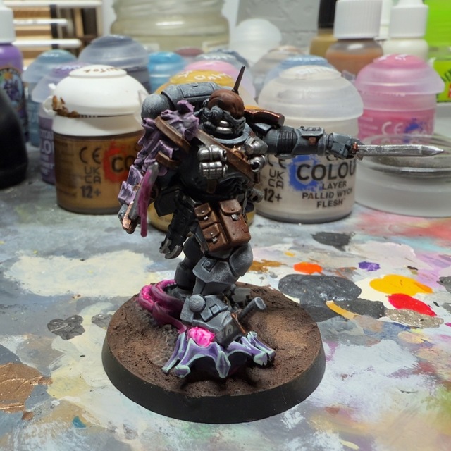 Close-up photograph of a Space Marine Phobos lieutenant model on a painting desk. The Tyranid shoulder pad and tyranid he’s standing on have a dark purple base with high contrast bright edge highlighting for a cyberpunk effect