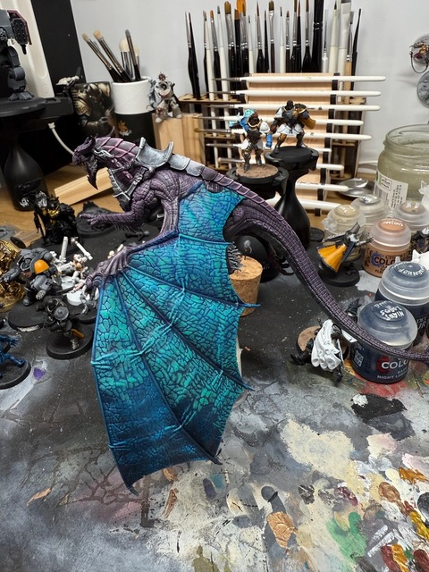 Photograph of a dragon model during painting. The wing has a blue base, with layers of blue-green highlighting applied to each individual scale. The final fifth or so of the wing is still to be highlighted, showing the deeper blue base colour