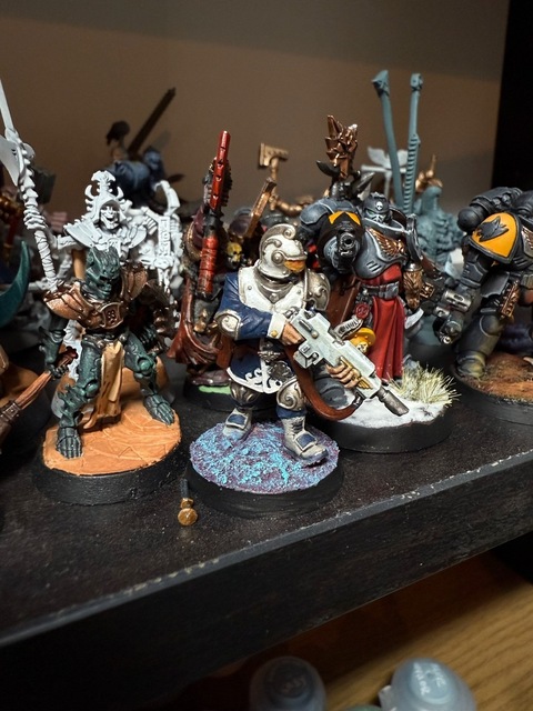 Photograph of a Voidsman model close up, standing on a black shelf along with a number of other miniatures. The base has a purple-ish ground with bright light-blue highlighting, while the models coat is dark blue and white, with a warm silver-ish metallic to the helmet and other areas
