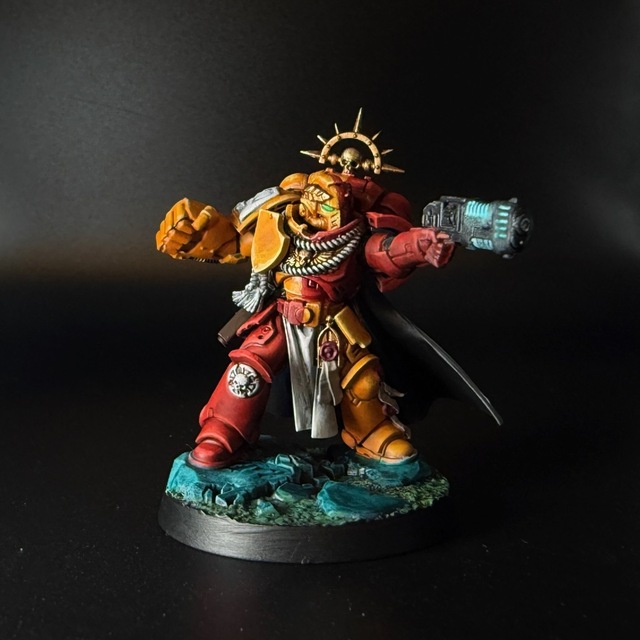 Photo of a painted space marine captain model on a black background. The miniature has the red and yellow quartered scheme of the Howling Griffons, with some edge highlighting on the armour and a blue-green scheme on the base rocks