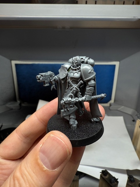 Photograph of hand holding a grey plastic miniature. It’s a space marine converted from a Stormcase Lord-Veritant model, with space marine shoulder pads and arms, and a power pack, plus some space wolf daggers on the belt. The left hand is holding a power axe while the right hand has a bolt pistol