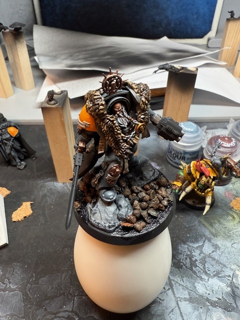 Photo of the terminator model on a white painting handle, from in front and above. The armour is a dark grey, with the wolf pelt picked out in some basic. detail and the head showing more detail and contrast. Some smaller ‘pebbles’ have been glued to the base in amongst the larger rocks