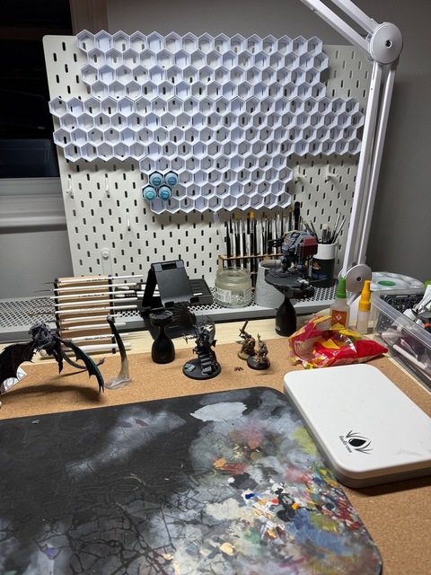 Photograph of a painting desk. A white pegboard is mounted standing at the back of the desk, with a honeycomb-like structure of hexagon-shaped white plastic paint pot holders. There are paint pots in a handful of the slots, pointing lid-out horizontally.