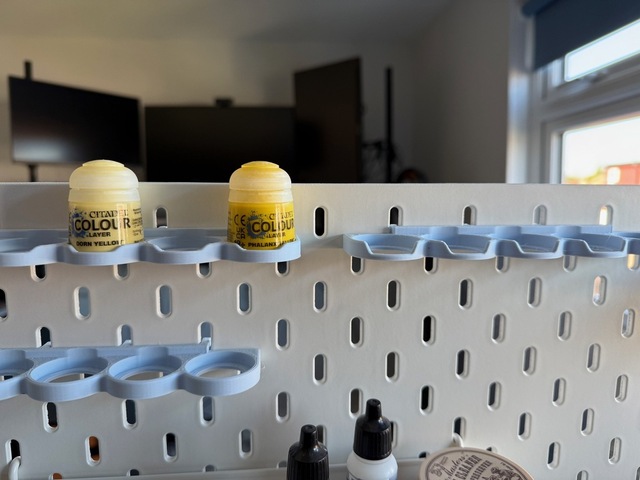 Photo looking head-on at a peg board. There are three horizontal racks shaped to hold Citadel paint pots upright. Each is an iteration of the design, adding a notch to reveal the printed paint name at the bottom of the pots.