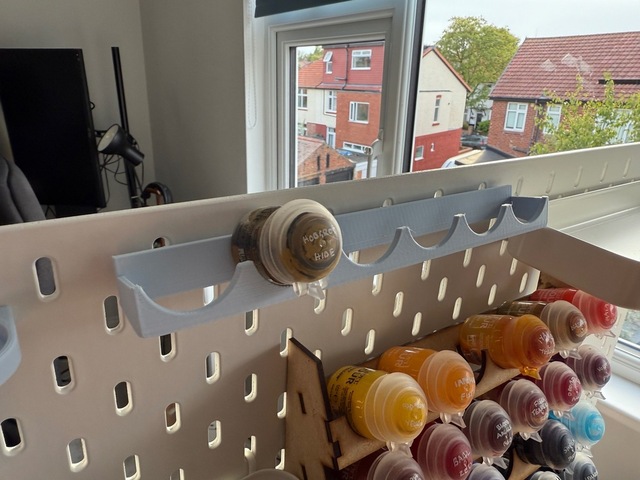 Photo of a pale blue 3D-printed paint rack mounted on a pegboard. It has a shallow, angled back plate across its width, with two arms to the front supporting a front plane with semi-circular cutouts to hold the shape of the paint pots