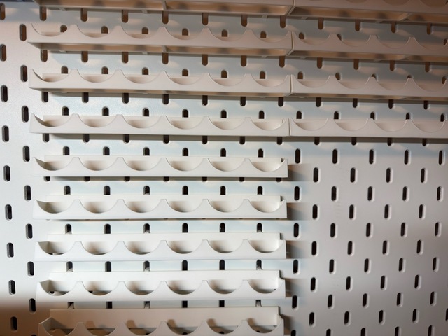 Photograph looking head-on at a pegboard with a couple of columns of 3d-printed paint racks, each with a row of semi-circular cutouts to hold a paint pot on its side.