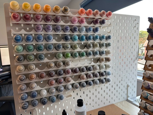 Photograph looking at a pegboard mounted to the side of a desk, with many rows of paint pots on racks in roughly colour wheel order, starting at yellow top-right and working down to neutrals at the bottom