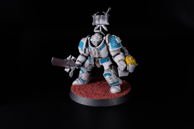 Photo on a stark black background of a Grey Knight terminator model, painted in a pale off-white, with runes a light-mid blue and much darker shade in the recesses
