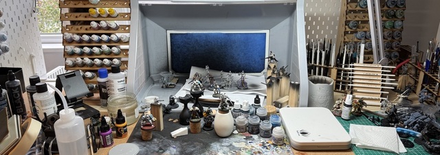 Photo of a desk covered in miniature painting materials. At the back is an airbrush booth, in front of which is a black 'texture mat' with many splashes of other coloured paints. Next to that a cutting mat and all around are many paints, miniatures and other bits of mess.
